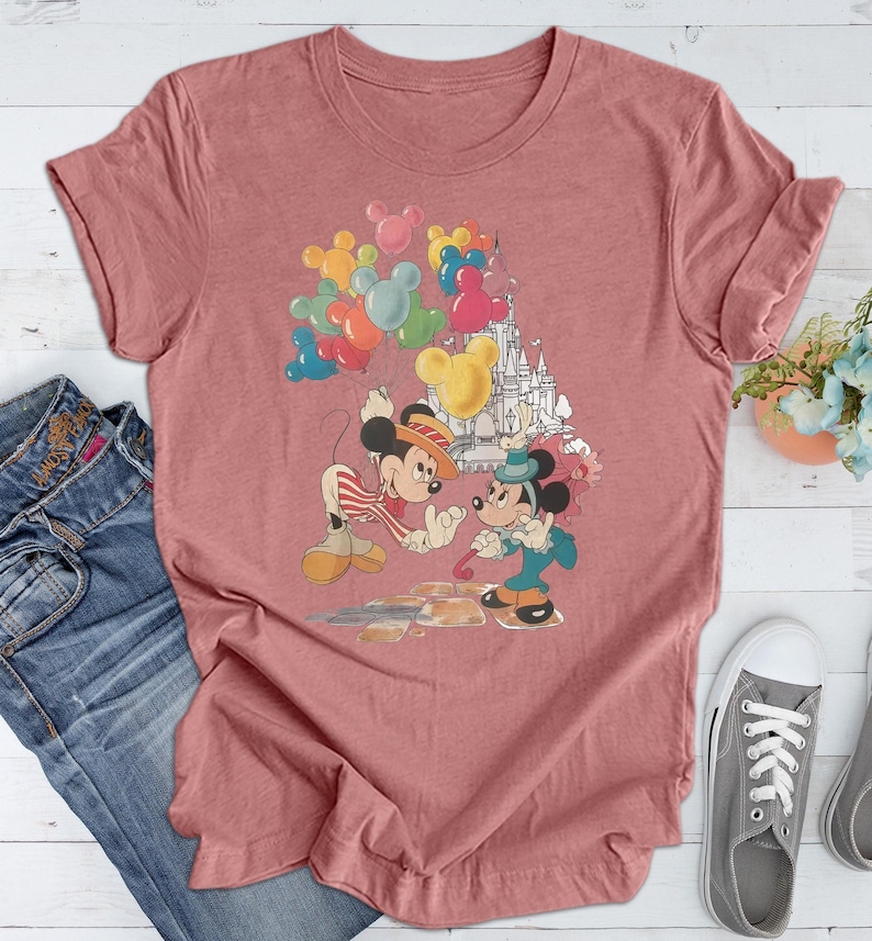 May include: A pink t-shirt with a vintage-style graphic of Mickey Mouse and Minnie Mouse holding balloons in front of a castle. The graphic is in shades of red, yellow, blue, and green.