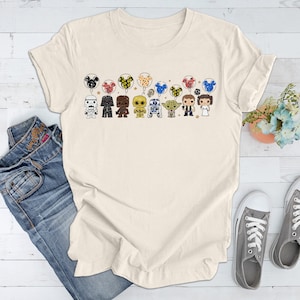 May include: A cream colored t-shirt with a graphic of Star Wars characters holding Mickey Mouse balloons. The characters include Darth Vader, Chewbacca, R2-D2, Yoda, Han Solo, Princess Leia, and a Stormtrooper.