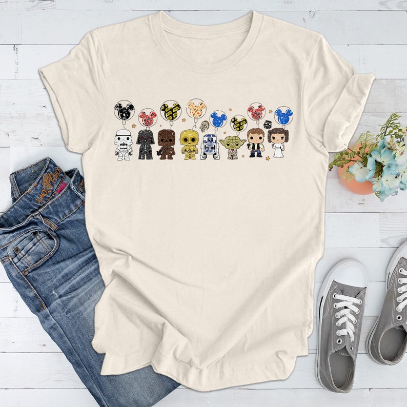 May include: A cream colored t-shirt with a graphic of Star Wars characters holding Mickey Mouse balloons. The characters include Darth Vader, Chewbacca, R2-D2, Yoda, Han Solo, Princess Leia, and a Stormtrooper.