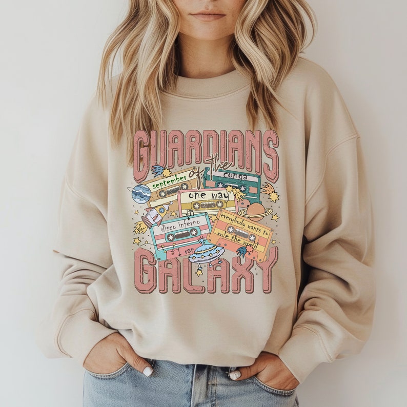 Peut inclure: Un sweat-shirt beige avec un imprim&eacute; graphique de cassettes audio et le texte "Guardians of the Galaxy". Le graphique est en rose, bleu et vert.