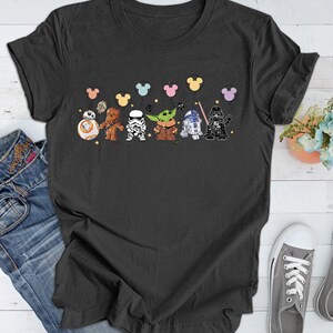 May include: A dark gray t-shirt with a graphic of Star Wars characters holding colorful balloons. The characters include Chewbacca, Darth Vader, R2-D2, a Stormtrooper, and Yoda.