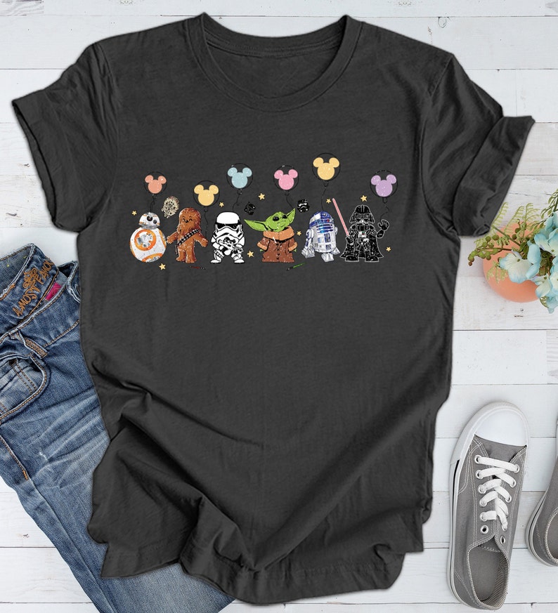 May include: A dark gray t-shirt with a graphic of Star Wars characters holding colorful balloons. The characters include Chewbacca, Darth Vader, R2-D2, a Stormtrooper, and Yoda.