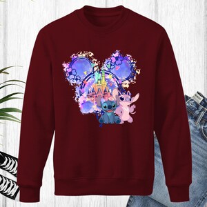 Puede incluir: Una sudadera granate con un gr&aacute;fico colorido de Stitch y Angel de Lilo & Stitch, con un castillo y un dise&ntilde;o de orejas de Mickey Mouse. El gr&aacute;fico incluye tonos de azul, rosa y morado. La sudadera se combina con unos vaqueros azules y zapatillas negras.