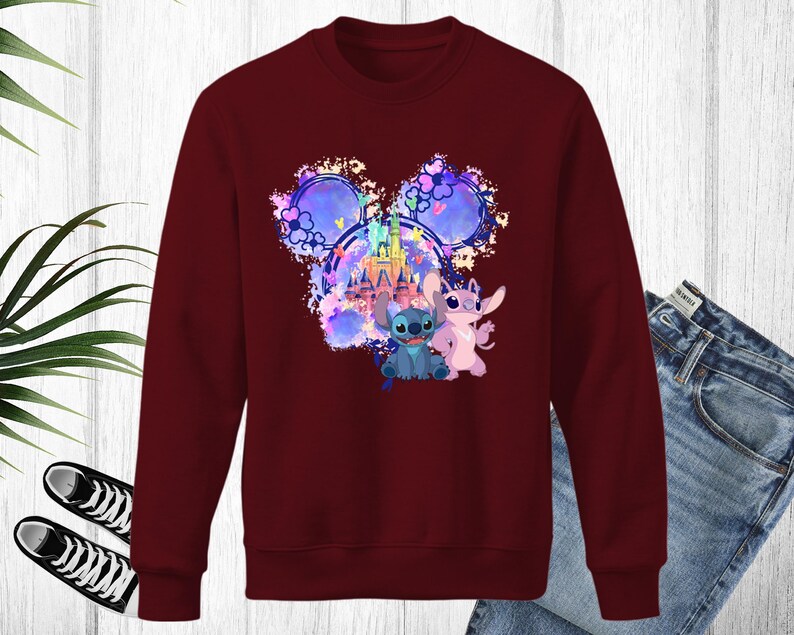 Puede incluir: Una sudadera granate con un gr&aacute;fico colorido de Stitch y Angel de Lilo & Stitch, con un castillo y un dise&ntilde;o de orejas de Mickey Mouse. El gr&aacute;fico incluye tonos de azul, rosa y morado. La sudadera se combina con unos vaqueros azules y zapatillas negras.