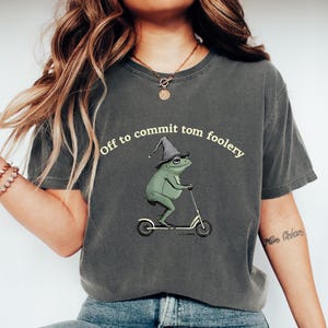 May include: Dark gray t-shirt featuring a whimsical illustration of a green frog wearing a wizard hat and riding a scooter. The text "Off to commit tom foolery" is arched above the frog. The shirt is a comfortable, casual style.