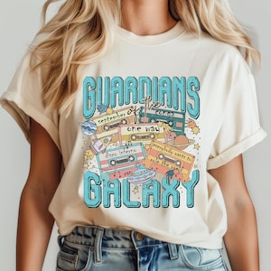 Peut inclure: Un t-shirt cr&egrave;me avec un motif graphique bleu et blanc qui dit "Guardians of the Galaxy" sur le th&egrave;me des cassettes audio r&eacute;tro. Le graphique comprend les mots "September", "One Way", "Disco Inferno", "Everybody Wants to Rule the World", et "I Ran".