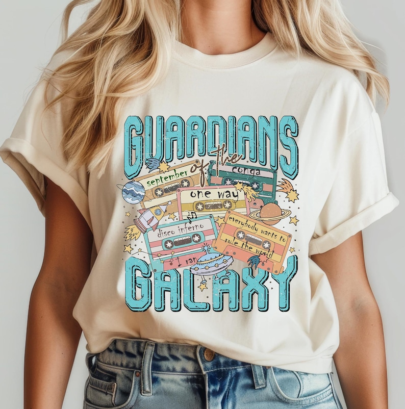 Peut inclure: Un t-shirt cr&egrave;me avec un motif graphique bleu et blanc qui dit "Guardians of the Galaxy" sur le th&egrave;me des cassettes audio r&eacute;tro. Le graphique comprend les mots "September", "One Way", "Disco Inferno", "Everybody Wants to Rule the World", et "I Ran".