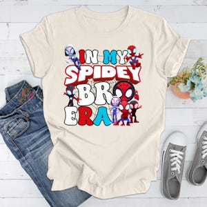 Puede incluir: Una camiseta beige con un dise&ntilde;o gr&aacute;fico que presenta varios personajes de Spider-Man de dibujos animados y el texto "In My Spidey Bro Era".