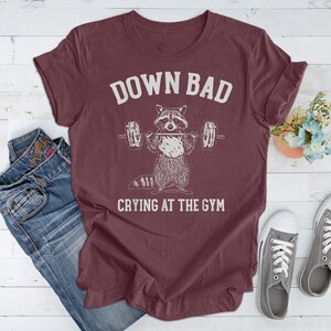 May include: Burgundy t-shirt with a white graphic of a raccoon lifting weights with the text "Down Bad Crying At The Gym".