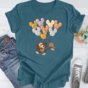 May include: Teal blue t-shirt with a cartoon dog and a cat holding onto a bunch of colorful Mickey Mouse balloons.