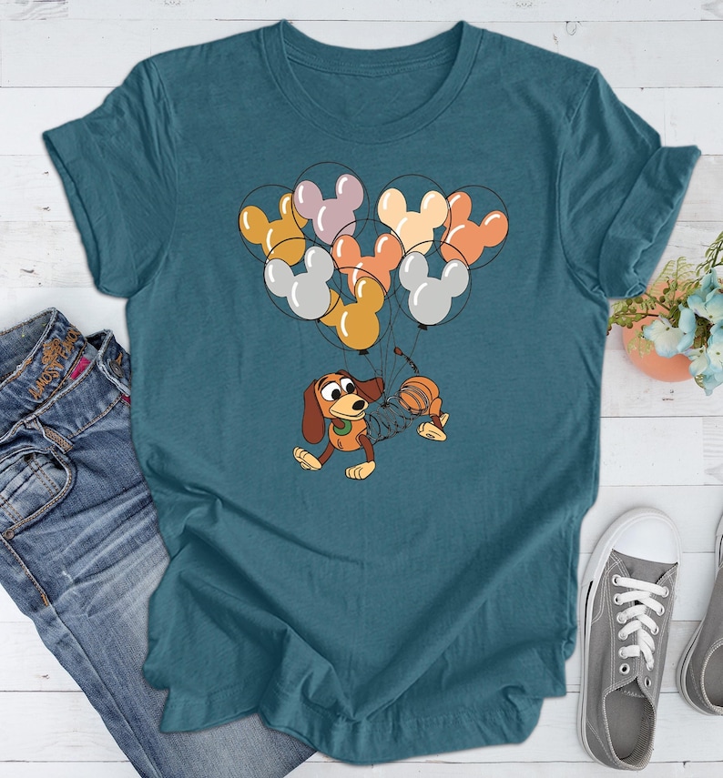 May include: Teal blue t-shirt with a cartoon dog and a cat holding onto a bunch of colorful Mickey Mouse balloons.