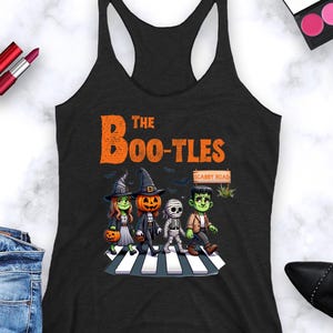 May include: Black tank top featuring a Halloween-themed graphic. The design shows four cartoon characters dressed as a witch, a jack-o'-lantern, a mummy, and Frankenstein crossing a crosswalk. The text "The Boo-Tles" is above the characters.