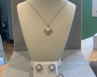 May include: A silver necklace with a square pendant featuring a pearl and small clear stones. The necklace is displayed on a white mannequin. A matching pair of earrings with a square design and a pearl are also shown.