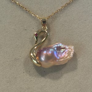 Può includere: Una collana color oro con un ciondolo a forma di cigno. Il cigno presenta un collo in metallo dorato lucido con una piccola pietra rossa come occhio e una grande perla barocca rosa chiaro iridescente che forma il corpo, appesa a una catena.