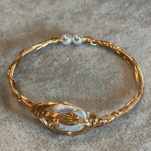 May include: A gold-toned wire-wrapped bracelet featuring a central oval-shaped iridescent stone encased in intricate metallic coils. The band is constructed from twisted gold wire and terminates at the opening with two small white spherical beads.