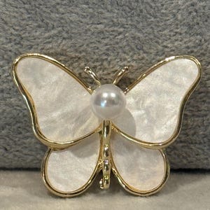 May include: A gold butterfly brooch featuring white, iridescent wings and a central pearl. The brooch has a delicate design, suitable for adding a touch of elegance to any outfit. The butterfly's wings have a subtle shimmer.