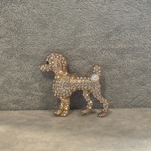 Puede incluir: Broche de caniche dorado, adornado con brillantes transparentes. El alfiler con forma de perro presenta un detalle de perla en la cola. Este broche decorativo está diseñado para añadir brillo a la ropa o los accesorios.