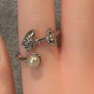 May include: A silver-toned open ring featuring a sculpted rose blossom on one end and a single round cream-coloured pearl on the other. The band is shaped like a slender rose stem with small leaves, creating a bypass style design that wraps around the finger.