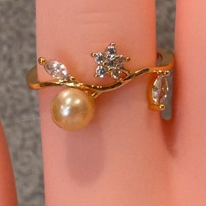 Könnte beinhalten: Ein goldfarbener Ring mit einer geschwungenen Ringschiene, die an eine Weinrebe erinnert. Das Design umfasst eine einzelne runde, cremefarbene Perle, eine kleine fünfblättrige Blume mit klaren Steinen und zwei marquise-geschliffene Steine.