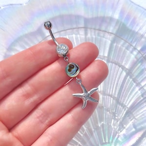 May include: A silver belly button ring with a curved barbell design. It features a clear gem at the top, a round abalone shell bead, and a starfish charm. The jewellery is designed for body piercing.