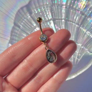 May include: Gold belly button ring with a clear gem and a teardrop-shaped dangle with a iridescent shell design.