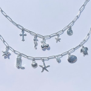 May include: Two silver charm necklaces featuring pendants like a cross, seahorse, cherries, starfish, spiral, star, cowboy boot, seashell, and turtle. Set against a white backdrop.