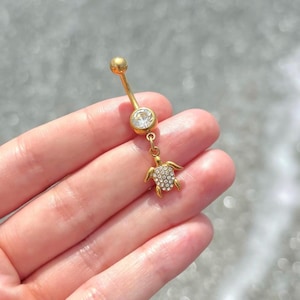 May include: Gold-coloured belly button ring with a clear gem and a small turtle charm. The turtle charm is encrusted with small, clear crystals. The jewellery is displayed against a blurred background of water.