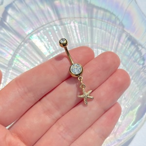 May include: Gold-coloured belly button ring with a dangling starfish charm. The ring features two clear, round gemstones and a small starfish pendant. The jewellery is displayed against a pearlescent background.