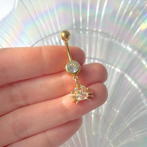 Peut inclure: Anneau de nombril doré avec une gemme ronde et transparente et un petit charme en forme de crabe. Le charme en crabe est orné de gemmes transparentes. Le bijou est tenu dans une main sur un fond nacré.