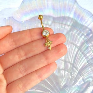 May include: Gold-colored belly button ring with a clear, round gem at the top and a small palm tree charm. The palm tree charm has a small, clear gem. The jewelry is held in a hand.