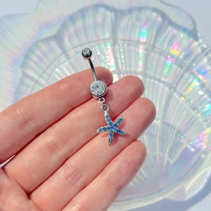 May include: A silver belly button ring with a clear gem at the top and a larger clear gem in the center. A blue rhinestone starfish charm hangs from the center gem. The jewelry is held in a hand.