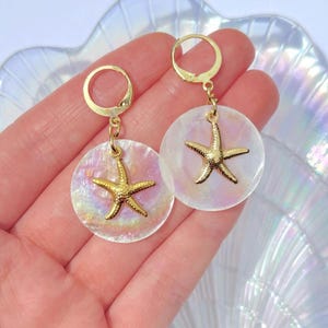 May include: Gold starfish earrings. Each earring has a gold starfish charm suspended from a gold hoop. The starfish hangs from a round, iridescent, mother-of-pearl disc. The earrings are designed for pierced ears.