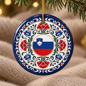 Op de afbeelding: Een rond keramisch ornament met de Sloveense vlag in het midden, omringd door een decoratieve rand van rode en blauwe bloemen met groene bladeren op een witte achtergrond, afgewerkt met een donkerblauwe rand en een gouden ophangkoord.