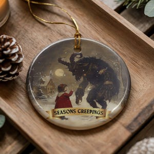 Puede incluir: Adorno navideño redondo con una escena invernal que presenta a un niño con abrigo rojo frente a una criatura oscura y grande. El adorno tiene una cuerda dorada para colgar y el texto "SEASONS CREEPINGS" en una pancarta.
