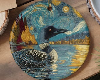 May include: A round ceramic ornament featuring a loon swimming on a lake. The artwork displays a swirling, starry night sky in shades of blue and yellow, with autumn-colored trees in the background. A gold string is attached to the top.