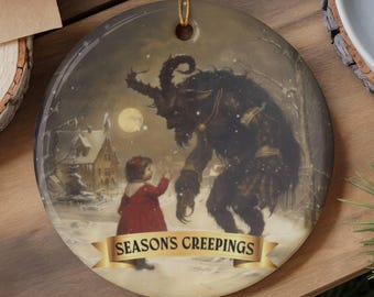 May include: A round ceramic ornament featuring a dark, horned creature standing in a snowy landscape with a child in a red coat. A gold banner at the bottom displays the text "SEASON'S CREEPINGS" in black lettering. The ornament hangs from a gold string.