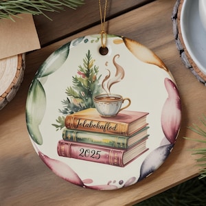 May include: Round ceramic Christmas ornament with a gold string. The ornament features a stack of books with a cup of coffee and a small Christmas tree. The books have the text "Jolabokafled" and "2025". The ornament has a watercolor floral design.