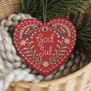 May include: A red heart-shaped Christmas ornament with the words "God Jul" in white script. The ornament is decorated with white flowers and green leaves. It hangs from a gold string and rests in a wicker basket.