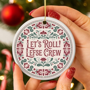 Op de afbeelding: Een ronde keramische kerstversiering met de tekst "Let's Roll! Lefse Crew" in bordeauxrood. De versiering heeft een witte achtergrond met een decoratieve rand van bordeauxrode en groene bloemelementen. Een gouden draad is bevestigd om op te hangen.