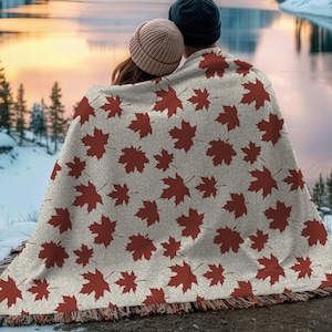 May include: A beige throw blanket with a pattern of red maple leaves covers two people, set against a scenic backdrop of a lake and snow-covered mountains. The blanket has a fringed edge, perfect for cozy outdoor moments.