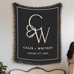 Peut inclure: Une couverture tissée noire avec une bordure et des franges blanches. La couverture affiche les initiales "C & W" entrelacées avec un motif de cœur, ainsi que les noms "Caleb + Whitney" et la date "2 août 2025".
