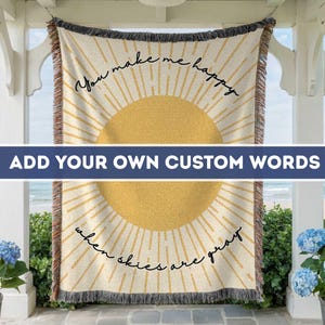 May include: Cream-colored woven throw blanket with a large yellow sun design and the phrases "You make me happy" and "when skies are gray" in black script. The blanket has a fringed edge and is displayed outdoors.
