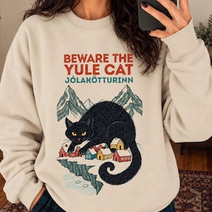 Op de afbeelding: Een beige sweater met ronde hals en een afbeelding van een grote zwarte kat die boven een klein dorpje met bergen op de achtergrond uitsteekt. De tekst BEWARE THE YULE CAT JÓLAKÖTTURINN staat in rode en blauwgroene letters erboven.
