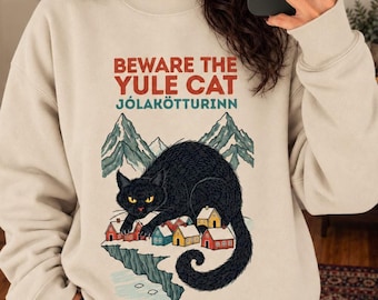 May include: A beige crewneck sweatshirt featuring a graphic of a large black cat looming over a small village with mountains in the background. The text reads BEWARE THE YULE CAT JÓLAKÖTTURINN in red and teal lettering above the illustration.