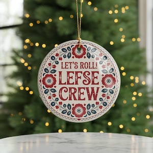 May include: Round ceramic ornament with a cream background, red floral designs, and the text "LET'S ROLL! LEFSE CREW" in red. The ornament is trimmed with a scalloped edge and a gold string for hanging. The background is a blurred Christmas tree.