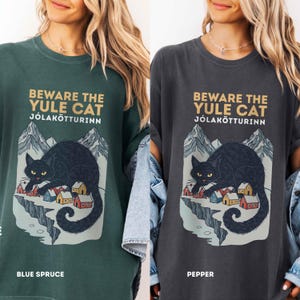 May include: A dark green and a charcoal gray t-shirt featuring a graphic print of a black cat looming over a small village with mountains in the background. The text reads BEWARE THE YULE CAT J&Oacute;LAK&Ouml;TTURINN. The shirts are labeled Blue Spruce and Pepper.