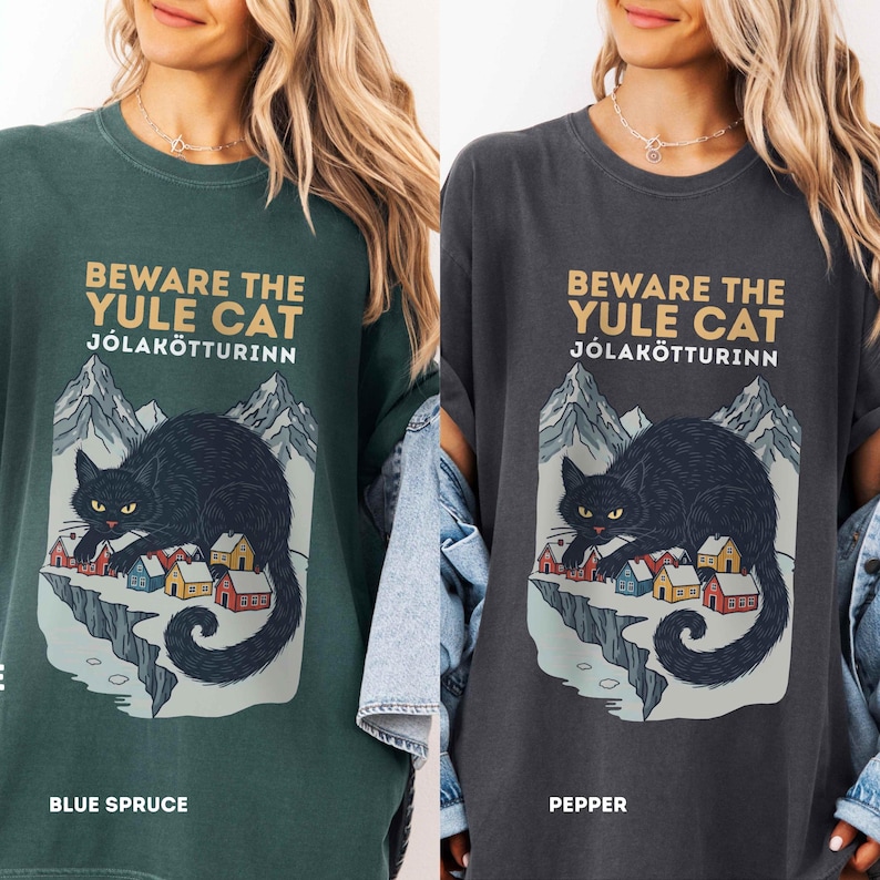May include: A dark green and a charcoal gray t-shirt featuring a graphic print of a black cat looming over a small village with mountains in the background. The text reads BEWARE THE YULE CAT J&Oacute;LAK&Ouml;TTURINN. The shirts are labeled Blue Spruce and Pepper.