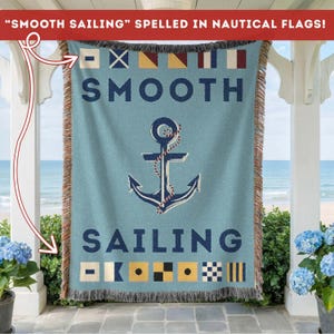 Puede incluir: Una manta tejida azul con las palabras "SMOOTH SAILING" y un diseño de ancla. Las palabras son en azul marino, y el ancla está detallada con una cuerda. La parte superior de la manta presenta banderas náuticas. La manta tiene un borde con flecos.