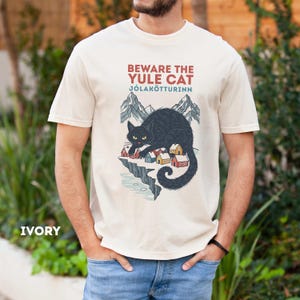 May include: An ivory t-shirt featuring a graphic of a large black cat looming over a small village with mountains in the background. The text reads BEWARE THE YULE CAT and J&Oacute;LAK&Ouml;TTURINN in red and blue lettering above the illustration.