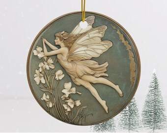 May include: A round ceramic ornament featuring a fairy with wings outstretched, flying over a cluster of white flowers. The fairy has long brown hair and a pink dress. The background is a muted green and brown.